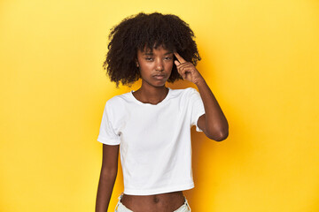 Teen girl in classic white T-shirt, yellow studio backdrop pointing temple with finger, thinking, focused on a task.