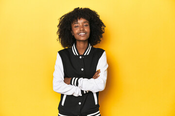 Teen girl in baseball jacket, yellow studio background who feels confident, crossing arms with...