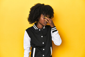 Teen girl in baseball jacket, yellow studio background having a head ache, touching front of the...