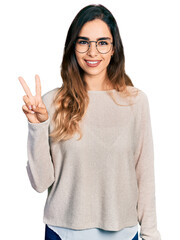 Beautiful hispanic woman wearing casual sweater and glasses smiling looking to the camera showing fingers doing victory sign. number two.