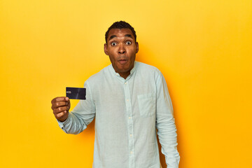 African American man with credit card, yellow studio, shrugs shoulders and open eyes confused.