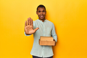 African American man with food boxes, yellow studio, standing with outstretched hand showing stop...