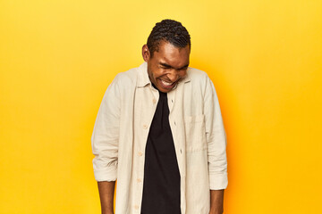 African American man with shirt, yellow studio, laughs and closes eyes, feels relaxed and happy.