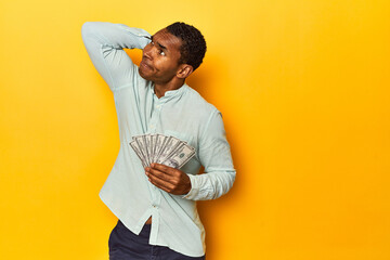 African American man with dollar bills, yellow studio, touching back of head, thinking and making a...