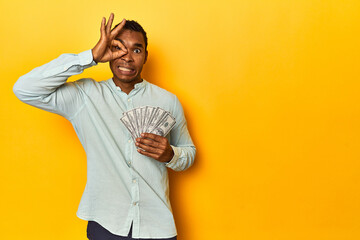 African American man with dollar bills, yellow studio, excited keeping ok gesture on eye.