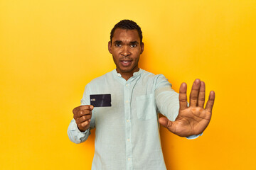 African American man with credit card, yellow studio, standing with outstretched hand showing stop...
