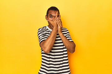 African American man in striped t-shirt, yellow studio, blink through fingers frightened and...