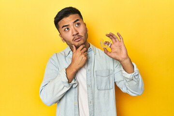 Asian man with invisible dental corrector, yellow studio backdrop looking sideways with doubtful...