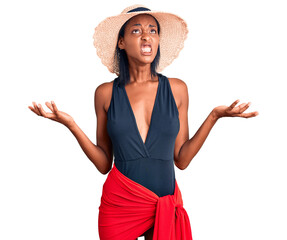 Young african american woman wearing swimsuit and summer hat crazy and mad shouting and yelling with aggressive expression and arms raised. frustration concept.