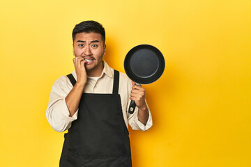 Asian chef holding a pan, yellow studio backdrop biting fingernails, nervous and very anxious.
