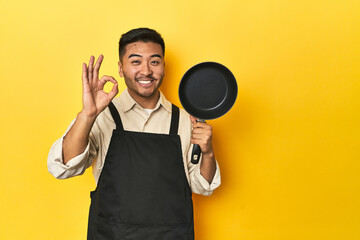 Asian chef holding a pan, yellow studio backdrop cheerful and confident showing ok gesture.