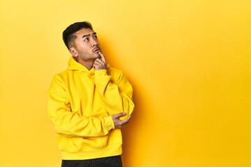 Bold Asian man in yellow hoodie on yellow studio looking sideways with doubtful and skeptical expression. - obrazy, fototapety, plakaty