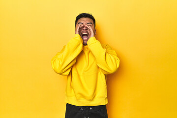 Bold Asian man in yellow hoodie on yellow studio shouting excited to front.