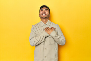 Asian man in stylish gray shirt on yellow studio laughing keeping hands on heart, concept of...