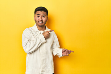 Asian man in crisp white shirt on yellow studio shocked pointing with index fingers to a copy space.