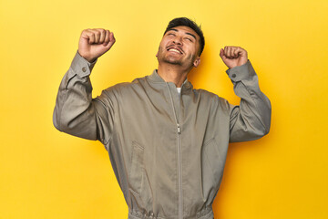 Stylish Asian man in gray jacket on yellow studio celebrating a special day, jumps and raise arms...
