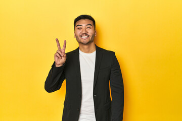 Asian man in black business jacket on yellow studio showing victory sign and smiling broadly.