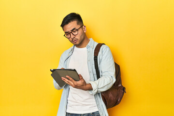 Asian student with backpack using tablet on yellow studio background.