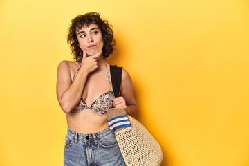 Caucasian woman in bikini with beach bag, studio looking sideways with doubtful and skeptical...