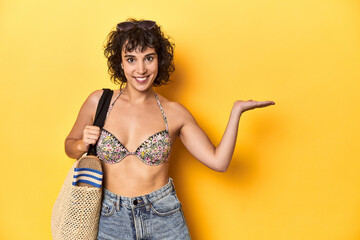 Caucasian woman in bikini with beach bag, studio showing a copy space on a palm and holding another hand on waist.