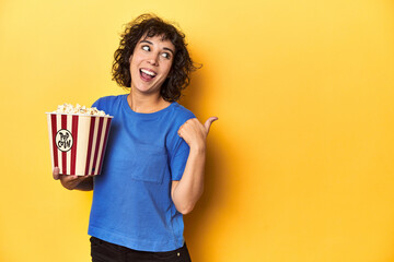 Curly-haired woman with popcorn for movie, studio points with thumb finger away, laughing and...