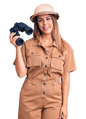 Young beautiful woman wearing explorer hat holding binoculars looking positive and happy standing and smiling with a confident smile showing teeth