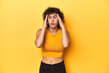 Curly-haired Caucasian woman in yellow top touching temples and having headache.
