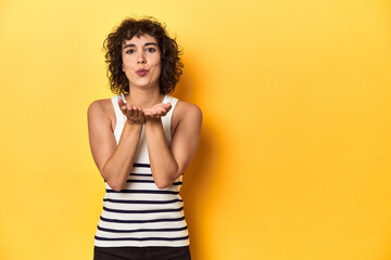 Caucasian curly-haired woman in white tank-top folding lips and holding palms to send air kiss.