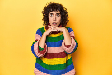 Curly-haired woman in multicolor sweatshirt praying for luck, amazed and opening mouth looking to front.