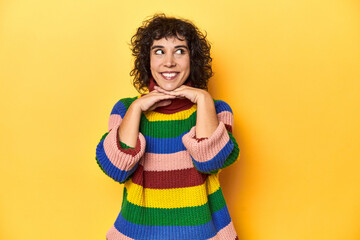 Curly-haired woman in multicolor sweatshirt keeps hands under chin, is looking happily aside.