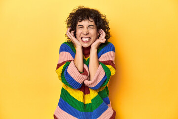 Curly-haired woman in multicolor sweatshirt covering ears with hands.
