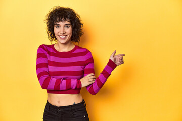 Caucasian curly-haired woman in pink striped-top smiling cheerfully pointing with forefinger away.