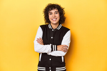 Curly-haired Caucasian woman in baseball jacket who feels confident, crossing arms with...