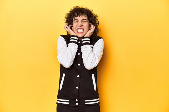 Curly-haired Caucasian woman in baseball jacket covering ears with hands.
