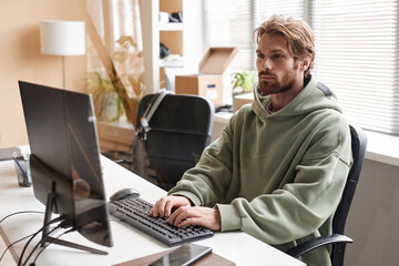 Portrait of young bearded software developer using computer and typing on keyboard while working in office, copy space