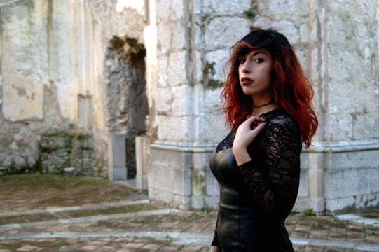 Amidst ruins, a red-haired dark beauty, half-bust framed, explores the mystery of an abandoned abbey with a haunting allure.