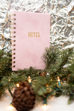 Vertical photo of a pink velor notepad on a Christmas background. Christmas mood, abstract background of crumpled foil and Christmas tree branches with a garland.