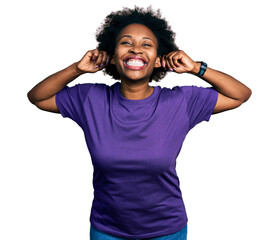 African american woman with afro hair wearing casual purple t shirt smiling pulling ears with...