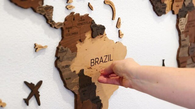A hand affixes a paper airplane-shaped pin to a textured wooden map of Brazil, symbolizing travel and adventure in South America.
