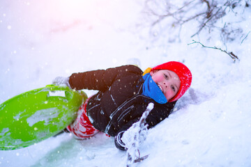 A little boy in a red hat and red pants lies on the snow of a snow slide with a round slide board. Tired and crawling up to slide down the hill