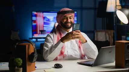 Arabic man at home in living room typing messages on smartphone. Muslim person holding mobile phone, enjoying relaxing leisure time talking with friends over online messaging app