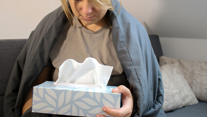 Sick caucasian woman sitting on sofa with blanket and holding a tissue box