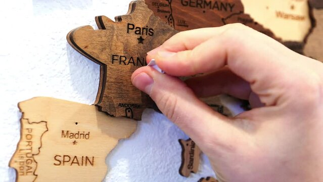 A hand carefully places a paper airplane-shaped pin over Paris on a wooden map of France, evoking themes of travel and exploration.