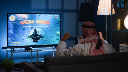 Arab gamer excited after winning game playing arcade space shooter on TV. Man spending time at home on gaming system, happy to finally complete singleplayer videogame mission