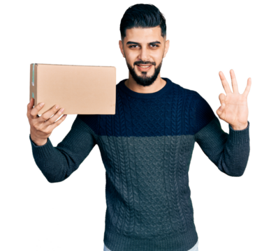 Young arab man with beard holding delivery package doing ok sign with fingers, smiling friendly gesturing excellent symbol