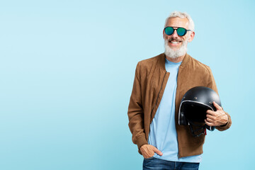 Cool handsome smiling mature man, gray haired bearded biker wearing stylish sunglasses, brown leather jacket holding motorcycle helmet isolated on blue background, copy space. Freedom concept