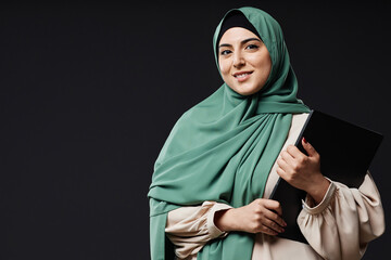 Waist up portrait of young Middle Eastern woman holding laptop and smiling at camera standing...