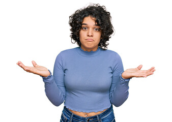 Young hispanic woman with curly hair wearing casual clothes clueless and confused expression with arms and hands raised. doubt concept.