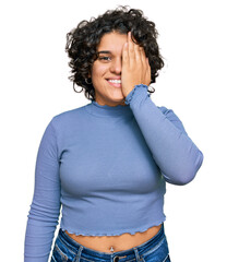 Young hispanic woman with curly hair wearing casual clothes covering one eye with hand, confident smile on face and surprise emotion.