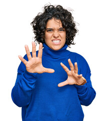 Young hispanic woman with curly hair wearing turtleneck sweater smiling funny doing claw gesture as...